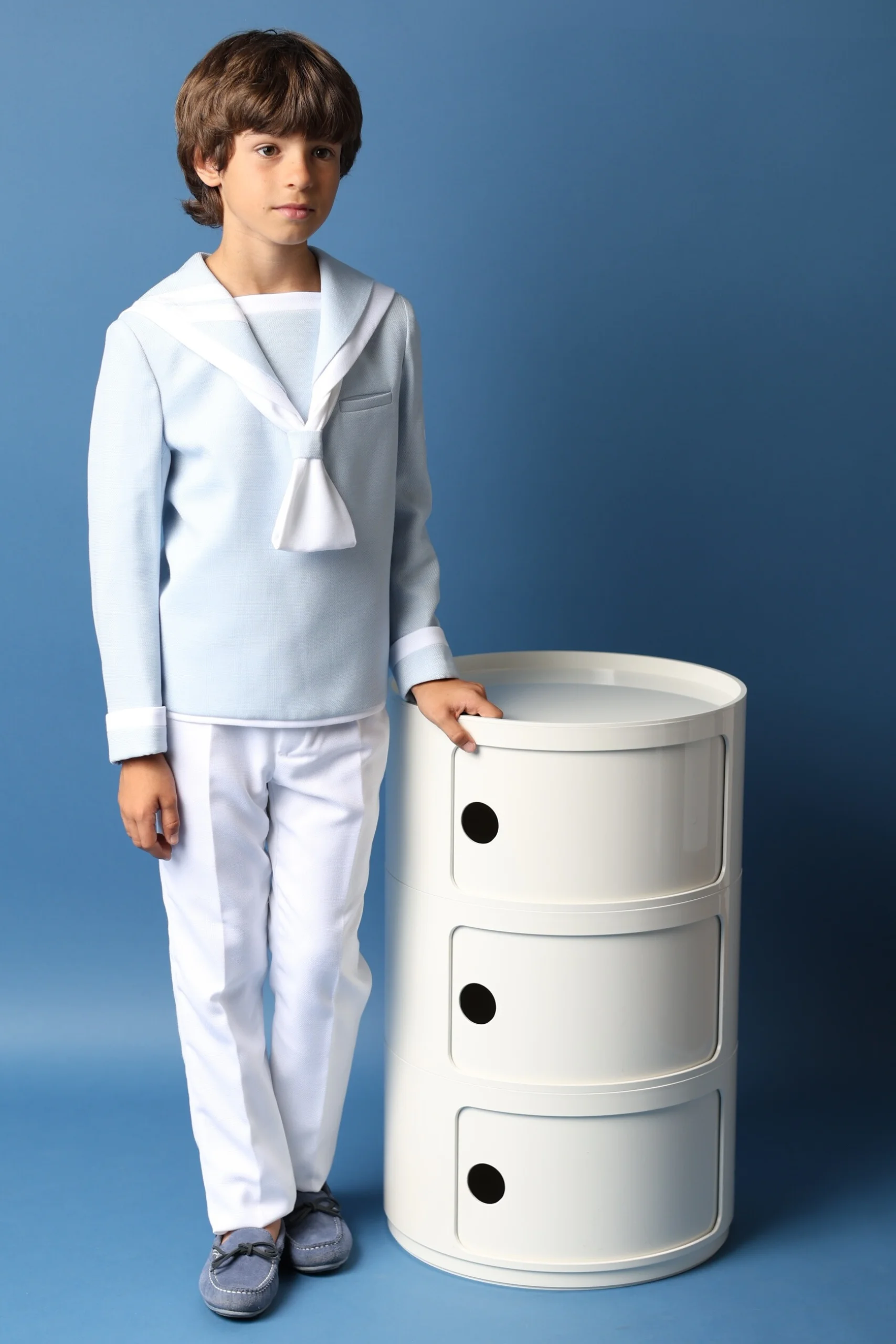 Traje de Comunión Marinero Niño 2025 en Blanco y Azul | Elegancia Atemporal Niño con traje de comunión marinero en blanco y azul claro, apoyado en un mueble moderno, ideal para ceremonias de comunión 2025.