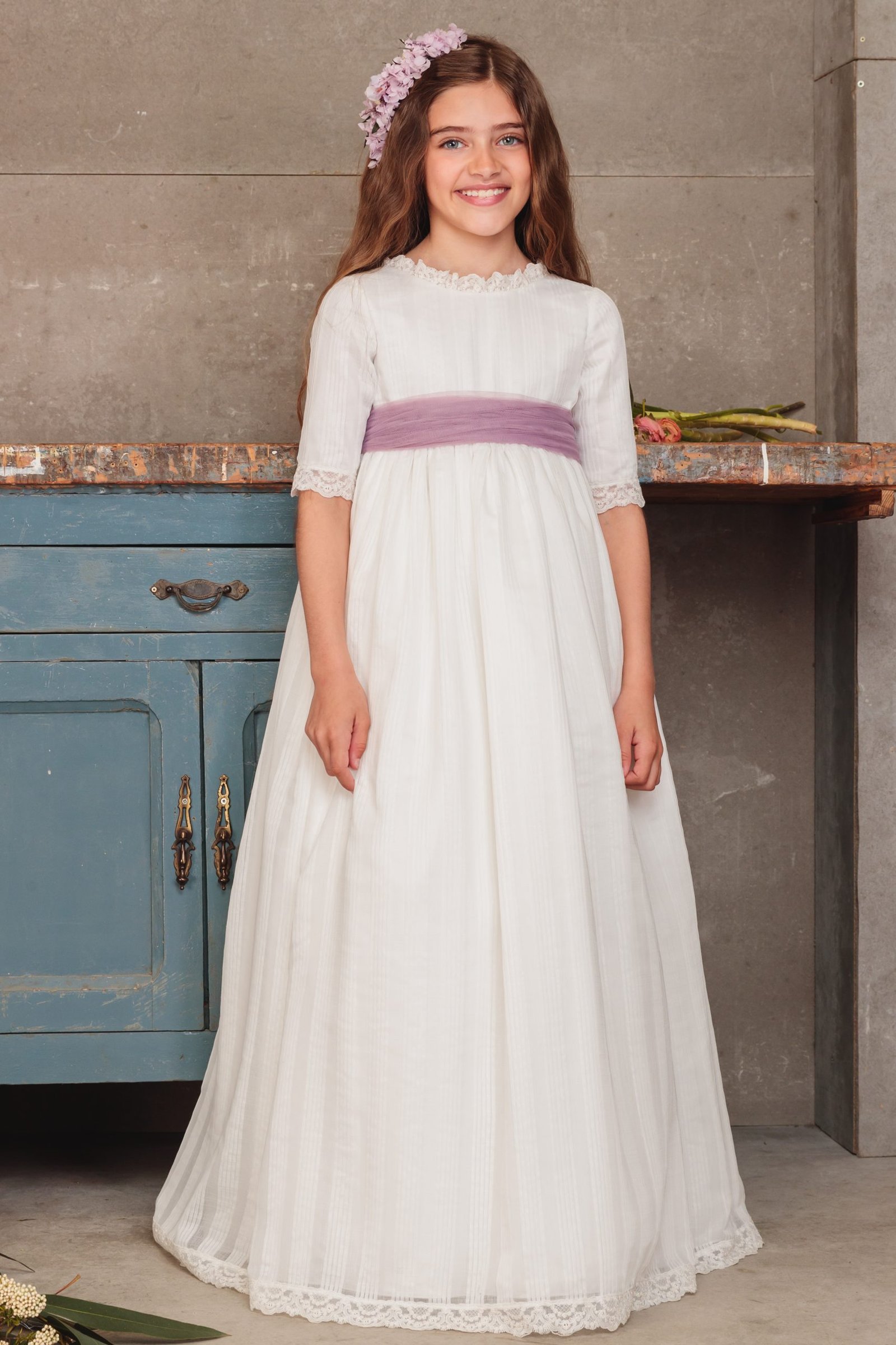 Vestido de comunión 2025 blanco con fajín lila, manga tres cuartos y corona de flores, disponible en MartaMolina.com